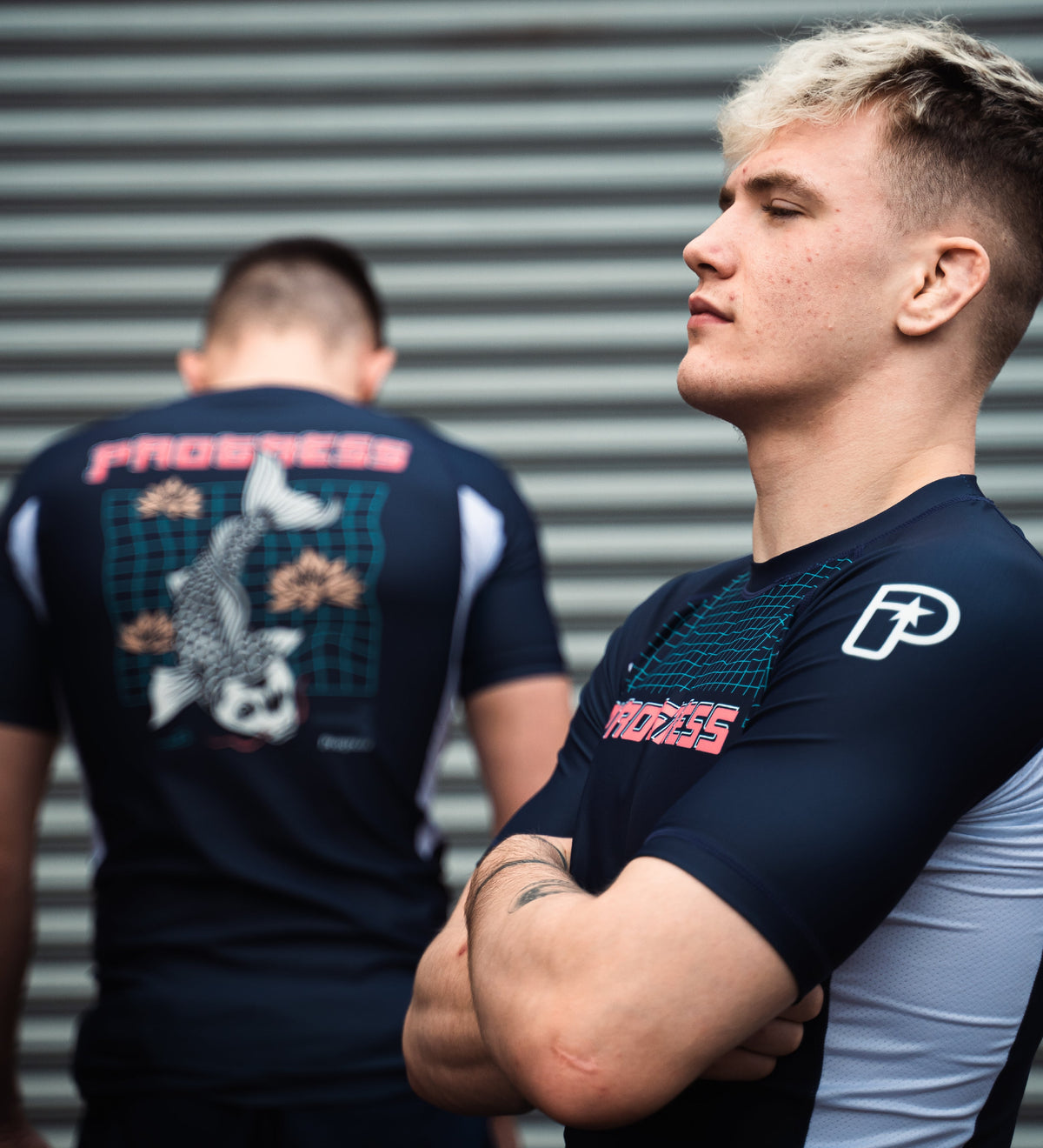 Owen Jones, a Brazilian Jiu-Jitsu athlete, stands with arms crossed wearing the navy blue Progress Jiu-Jitsu Unknown Koi rash guard, while another athlete in a matching rash guard  stands in the background against a metal backdrop.